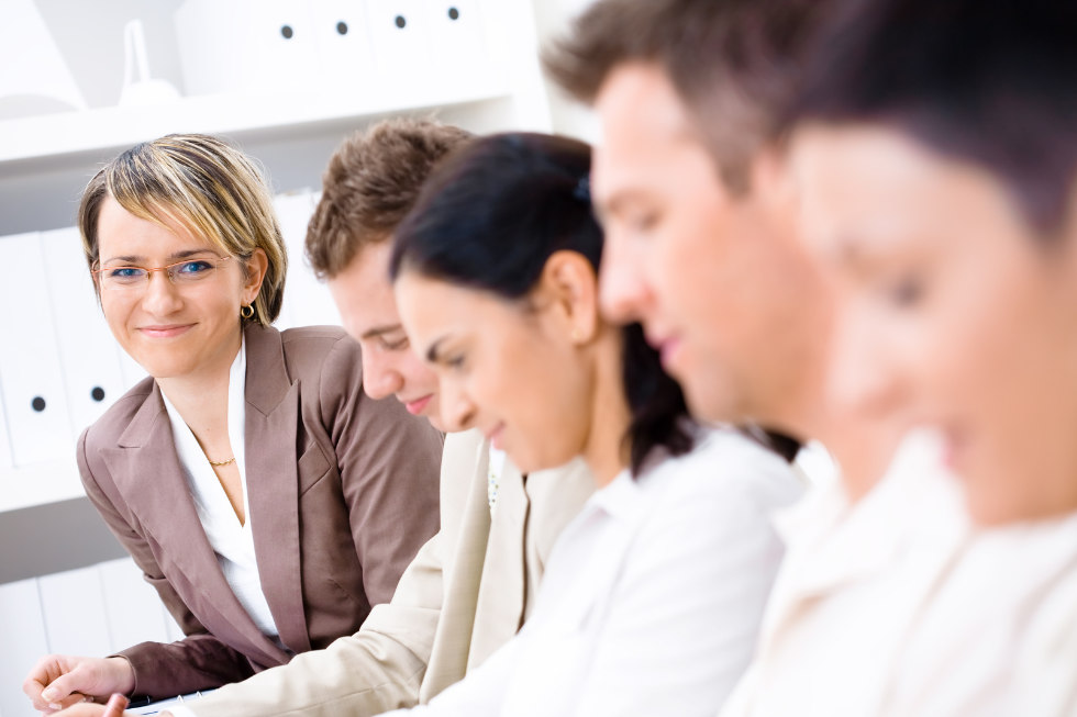 Vier Personen sitzen in einer Reihe in einem Büro im Cäsar Sprachinstitut; die Person links, die eine Brille und einen braunen Blazer trägt, lächelt und schaut in die Kamera.