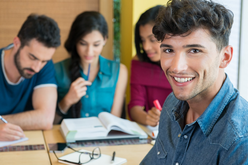 Ein junger Mann im Jeanshemd lächelt in die Kamera, während drei andere mit Büchern und Heften an einem Tisch im Hintergrund während eines Deutschkurses im Cäsar Sprachinstitut lernen.