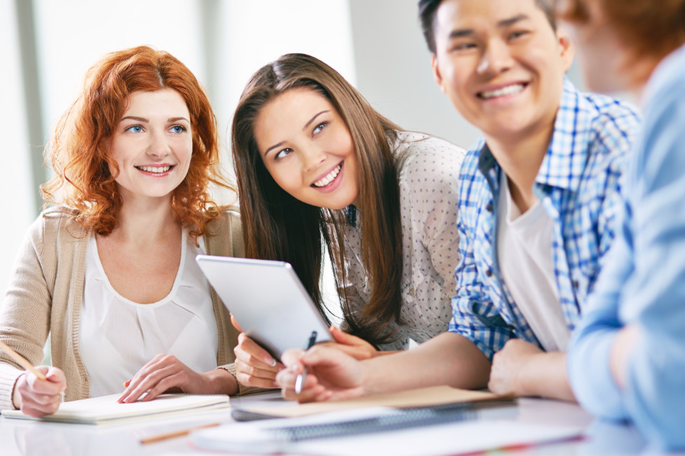 Vier junge Erwachsene sitzen lächelnd und plaudernd an einem Tisch, vor sich Notizbücher und ein Tablet, während sie sich auf ihren Deutschkurs im Cäsar Sprachinstitut vorbereiten.