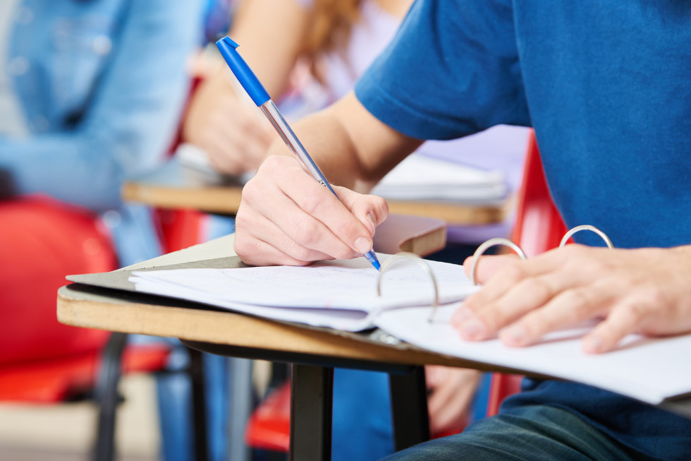 Eine Person schreibt mit einem blauen Stift auf Papier an einem Schreibtisch in einem Klassenzimmer während eines Deutschkurses, während andere Schüler im Hintergrund zu sehen sind.